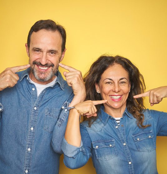 Beautiful middle age couple together standing over isolated yellow background smiling cheerful showing and pointing with fingers teeth and mouth. Dental health concept.
